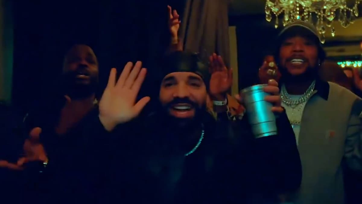 Still from the Drake Burning Bridges video: Drake in a black durag and black jacket waving at a handheld camera in a dim dining room, a man in a dark hoodie at left, a man in a grey jacket with chains raising a teal metal cup at right, a chandelier upper right