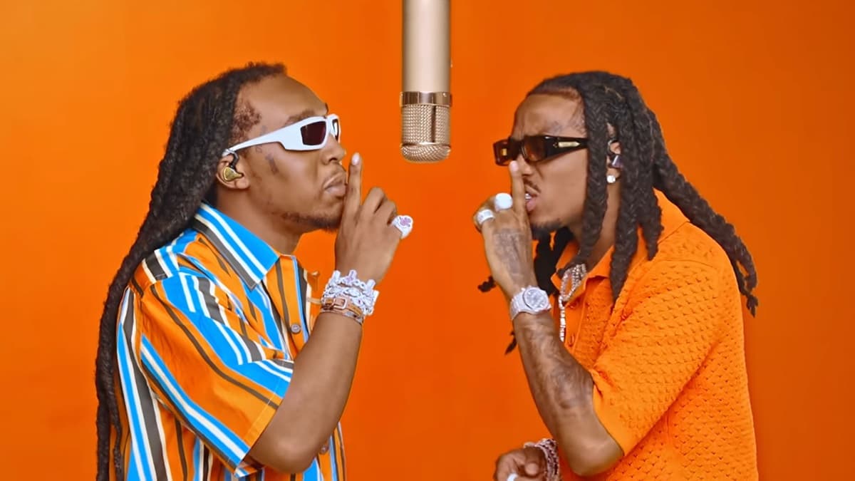 Still from Quavo and Takeoff's COLORS performance of Hotel Lobby: two rappers face each other in profile under a single hanging microphone against a solid orange backdrop, each holding a finger to his lips, one in a striped orange-and-blue shirt and white sunglasses, the other in an orange knit shirt and dark sunglasses
