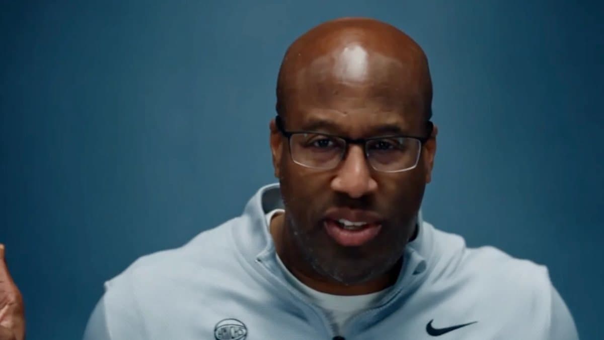 AI video frame: Mike Brown in glasses and a gray Nike Knicks quarter-zip against a blue backdrop, mid-gesture, in the shot that belongs to Chilli in the original
