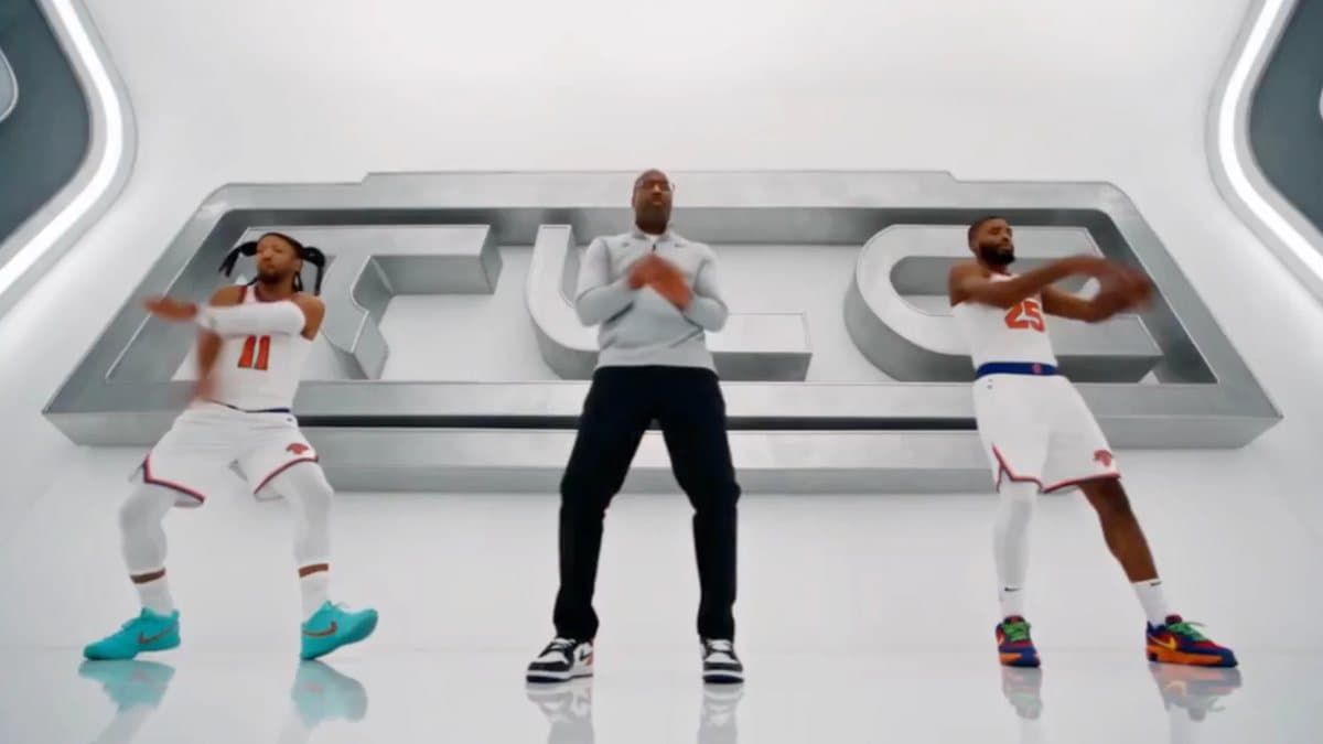 AI video frame: Jalen Brunson in braided pigtails, coach Mike Brown in a gray Knicks quarter-zip, and Mikal Bridges performing TLC's No Scrubs choreography in front of giant steel TLC letters