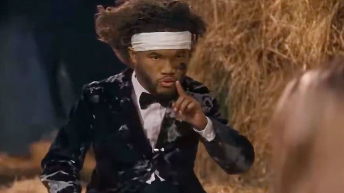 AI remix of the tiptoe barn clip: a man resembling Vikings quarterback Kyler Murray, with a large afro and a white headband, in a black tuxedo with white marbling, finger to his lips, hay bales behind him