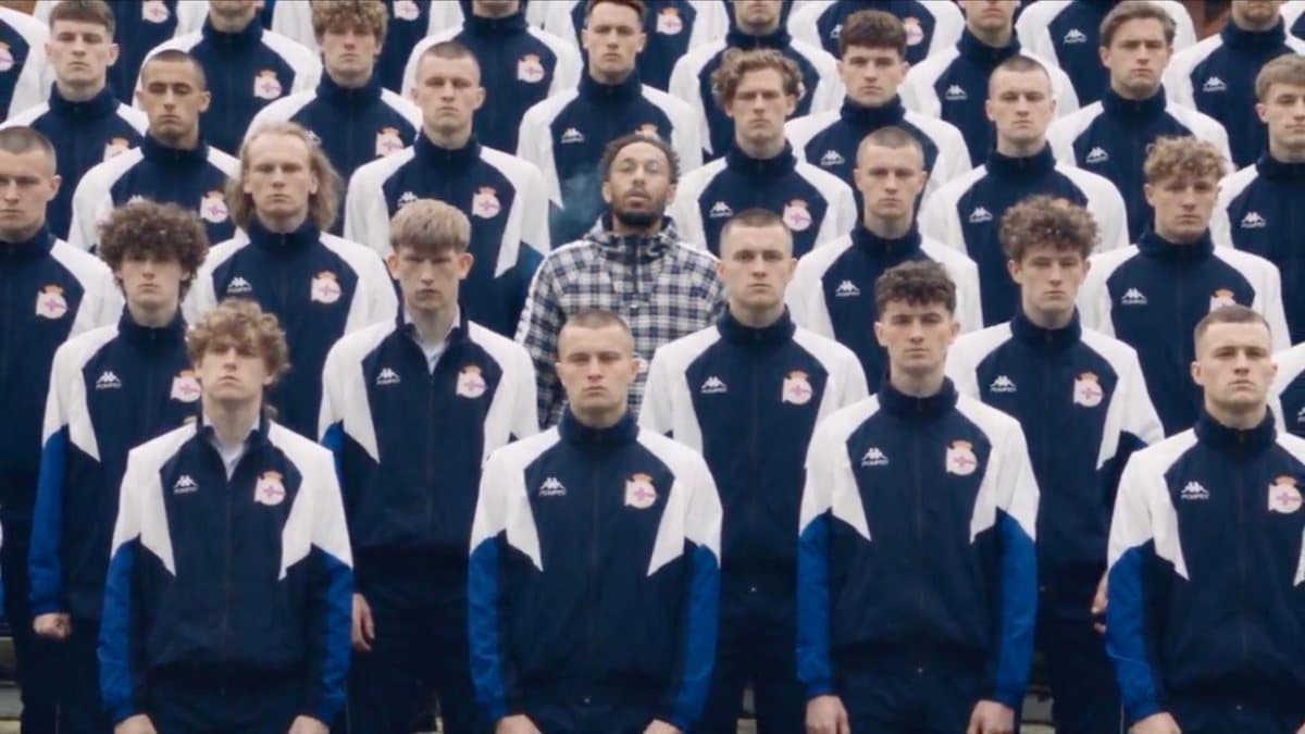 Still from an AI re-creation of the STORM crowd shot: rows of young men in navy, white, and blue Kappa tracksuits with a Deportivo crest, with footballer Pierre-Emerick Aubameyang in a plaid jacket standing motionless at the center, exhaling smoke