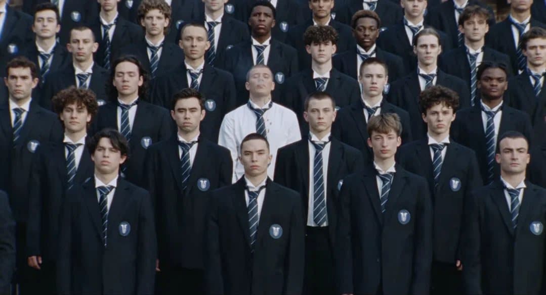 Still from GENER8ION's STORM starring Yung Lean: rows of schoolboys in dark crested blazers and striped ties assembled for a school photo, with one boy in a white shirt standing motionless at the center