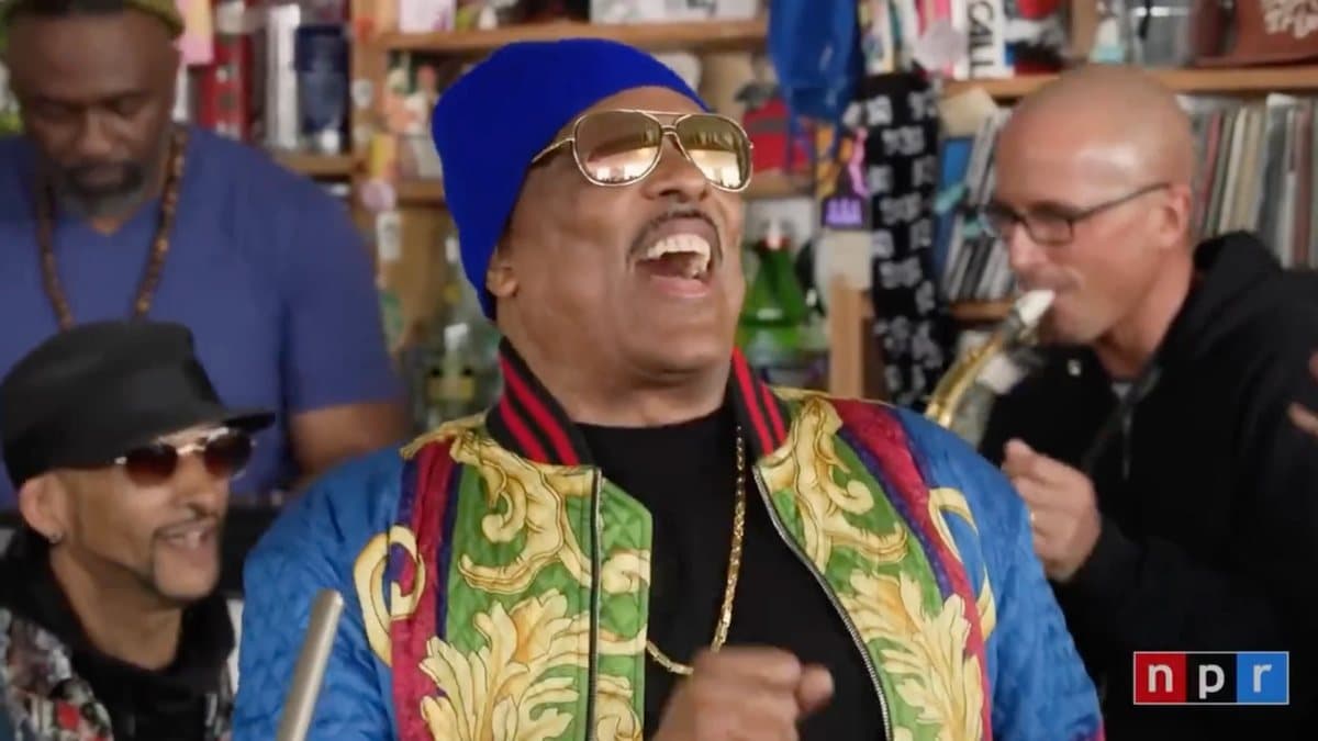 Charlie Wilson in a blue beanie, gold aviators, and a baroque-print jacket singing at the NPR Tiny Desk, saxophonist behind him