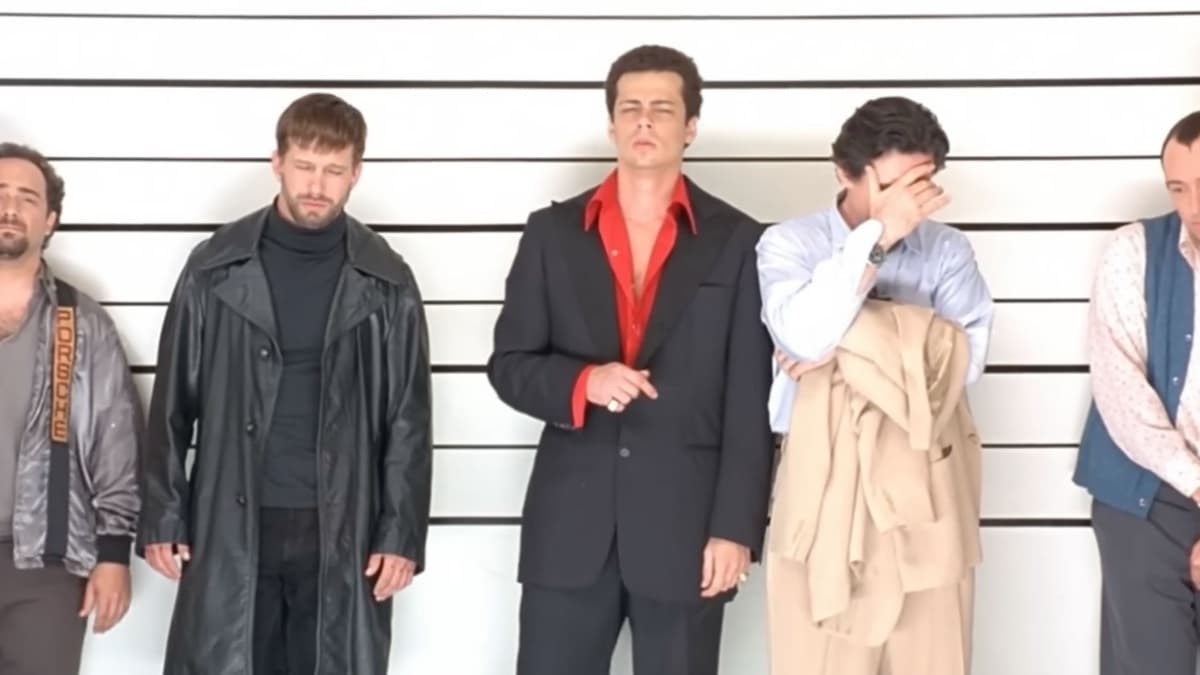 The original lineup from The Usual Suspects: five men against a white height-chart wall — a man in a satin jacket at left, Stephen Baldwin in a long leather coat, Benicio Del Toro in a black suit over a red shirt at center, Gabriel Byrne covering his face with his hand while holding a camel coat, and a man in a patterned shirt at right